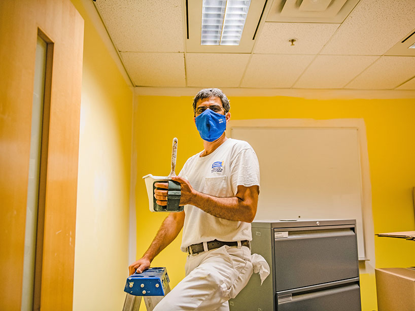 A man wearing a mask standing on a step ladder with a paint can and brush in his hand