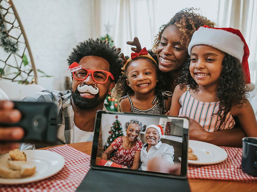 Two parents and their young daughters smile for a selfie with grandparents on another screen in front of them, also smiling