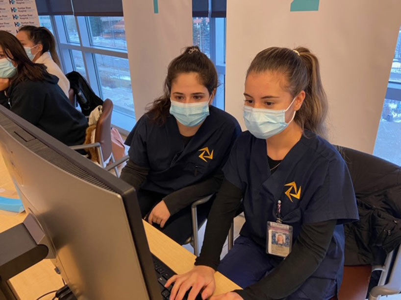 Two young women sit at a computer with masks on.