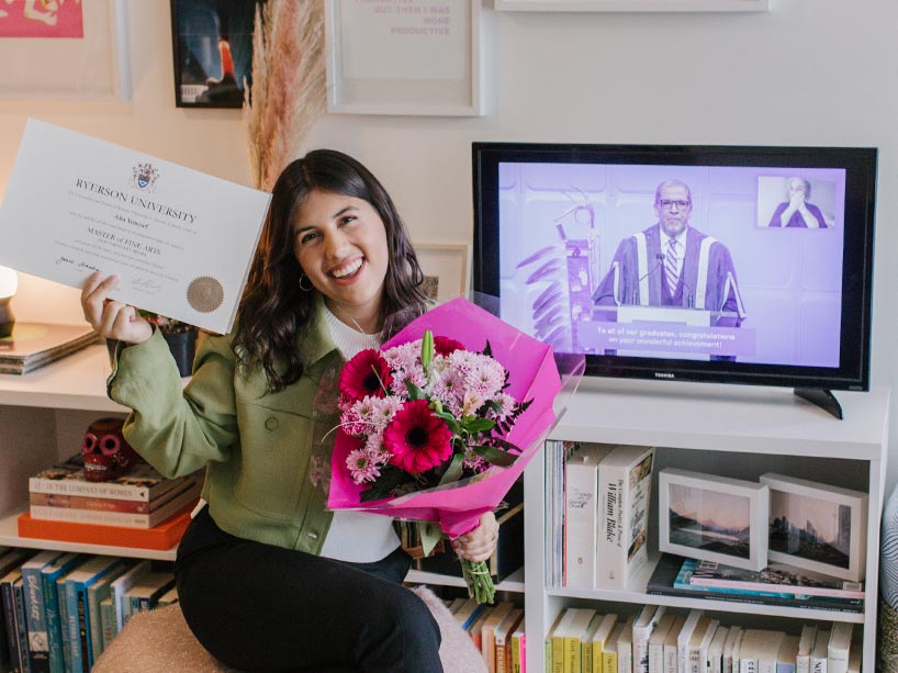 A young woman smiles while holding her newly conferred university degree, with flowers in her other hand, as a graduation ceremony plays on a screen behind her.