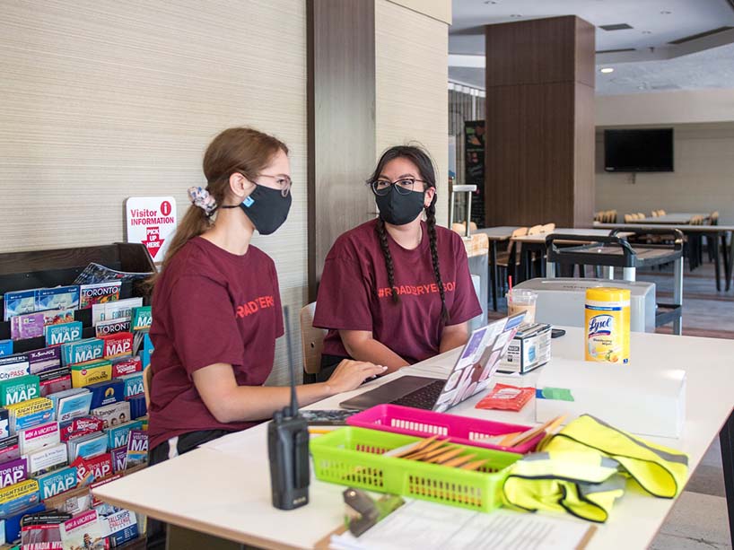 Photo of two people wearing masks behind a desk containing Lysol wipes, a computer and a walkie talkie.