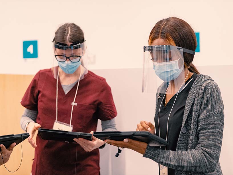 Two female nurses wearing masks and face shields stand with iPads in their hands.