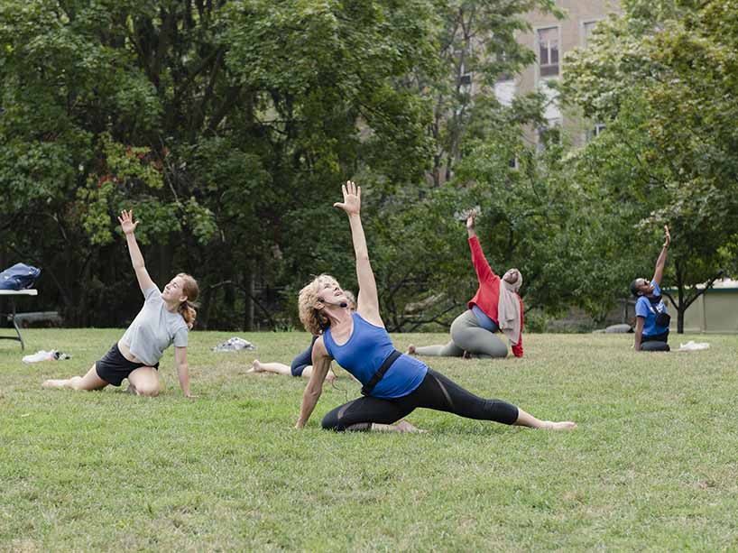 Three students stretching on a lawn behind a fitness trainer.