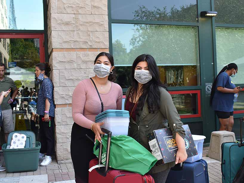 Two students wearing masks, holding their luggage and boxes.
