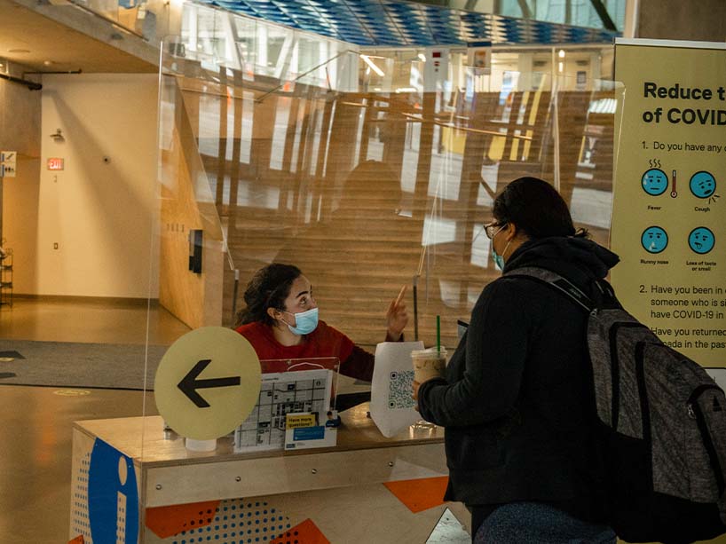 A student wearing a mask sits at a desk in the SLC giving info to a student wearing a mask standing on the other side of plexiglass