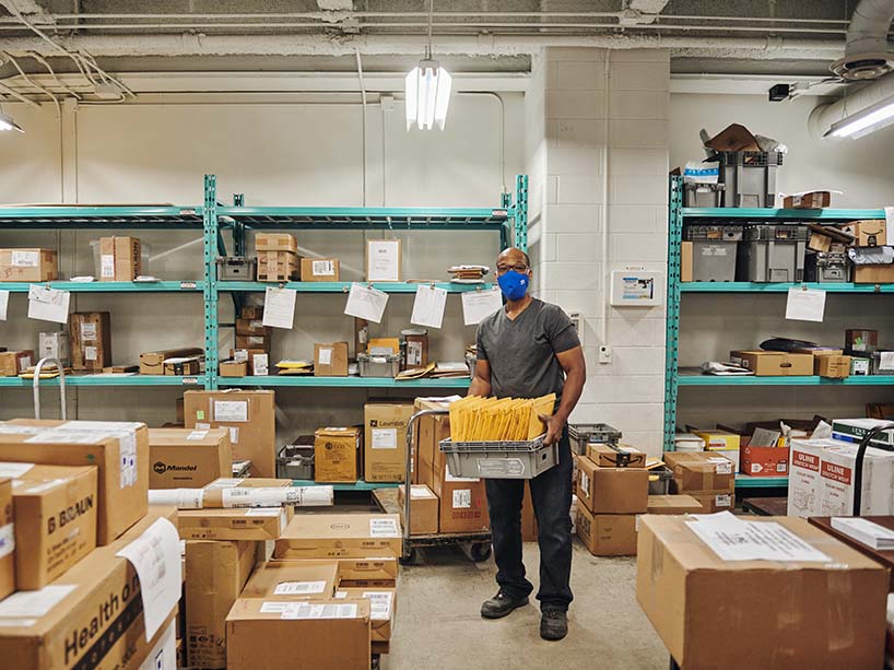 Howard O. Allen stands in the mail room carrying a box of small packages, surrounded by boxes for delivery.