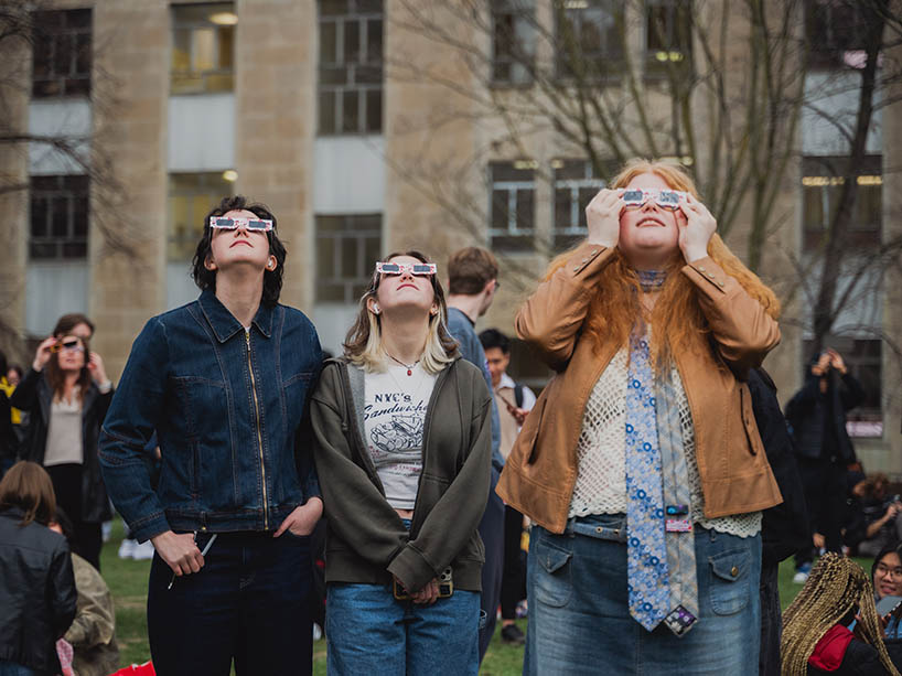 Three people look up at the sky with protective glasses on.
