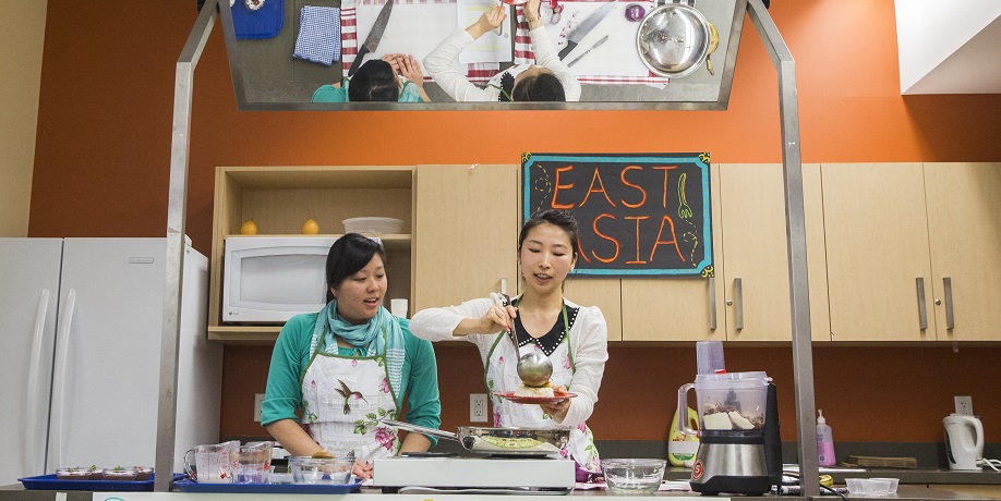 Nutrition students preparing East Asian food in kitchen for TMU food and cultural festival. A sign reading 'East Asia' hangs behind them.