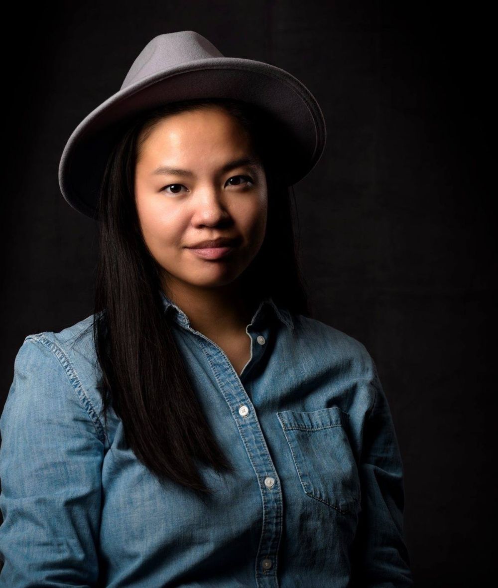 Eva smirks at the camera against a black background. She is wearing a large brimmed hat and a denim button up shirt