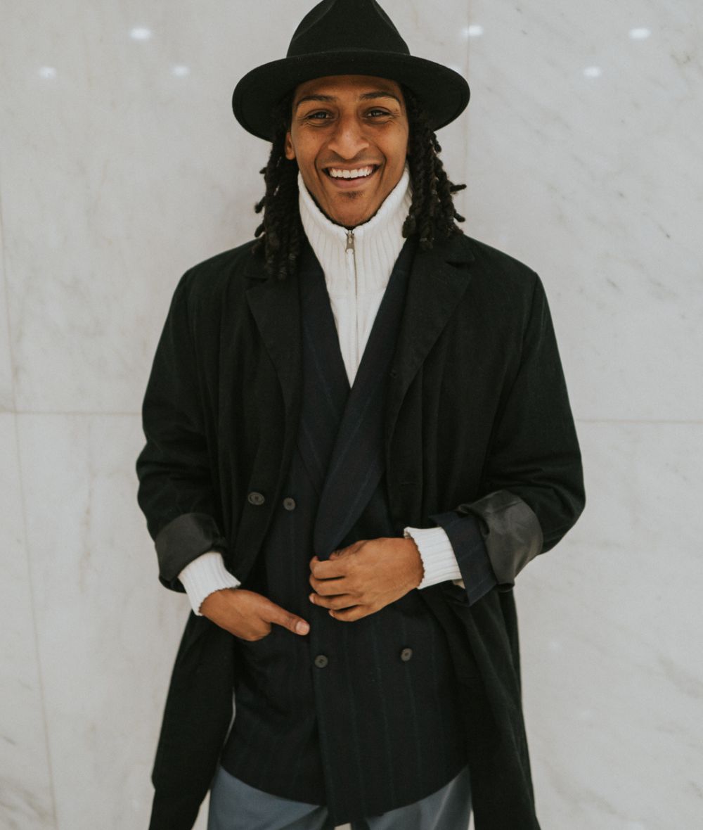 Raoul is standing in front of a grey background smiling. He is wearing a large brimmed hat and a woll coat, his hands are in his pockets. 