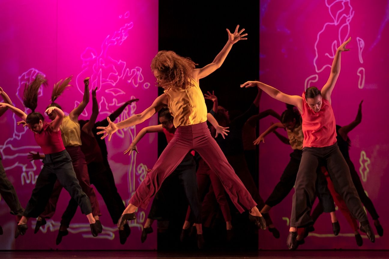 Dancers in jewel tones, jump with their arms and legs spread.