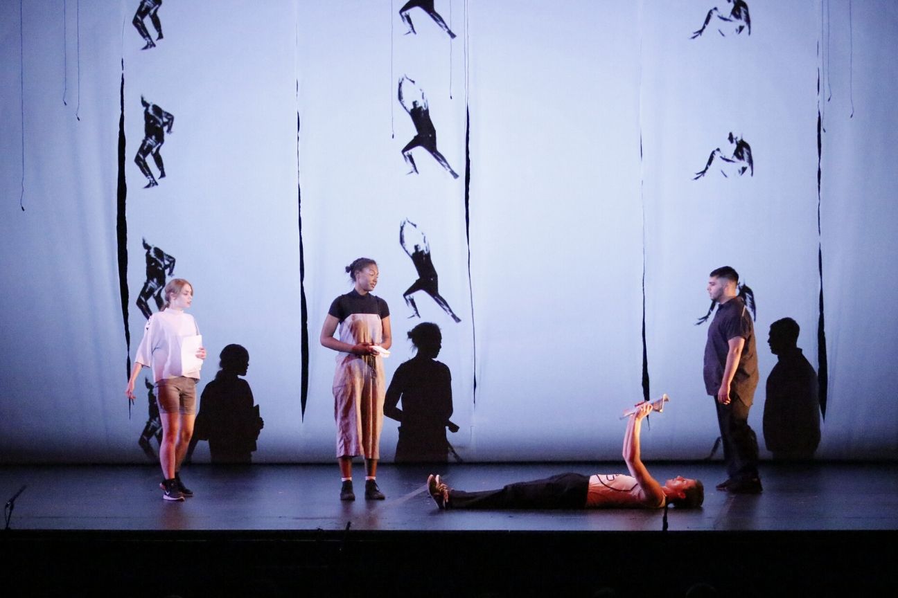 4 actors on stage, 1 lies down, projections of human shapes behind them.