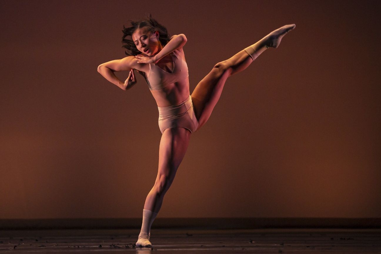 A dancer in a nude body suit lifts her leg very high.