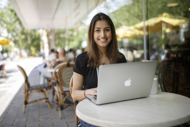 Student smiling at the camera while she works on her laptop outside at a Balzac's table.