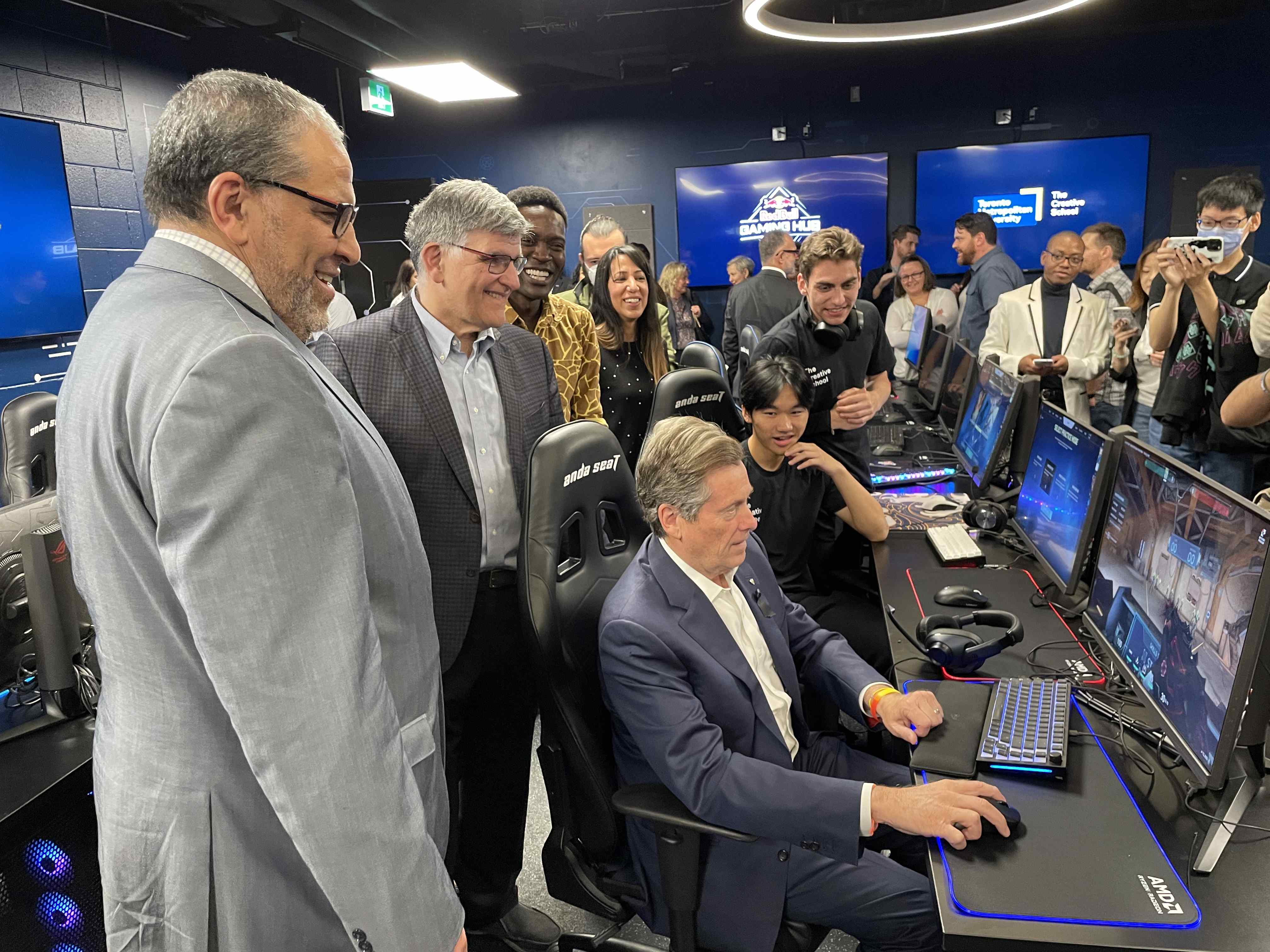 Mayor John Tory sits infront of a computer in the Red Bull Gaming Hub while President Lachemi and Dean Charles Fazon stand behind him
