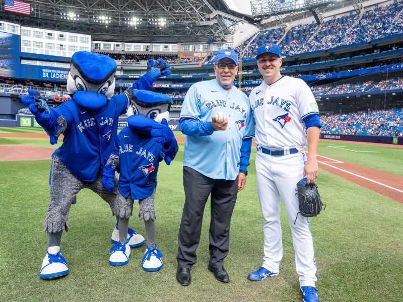 President Lachemi wearing a Toronto Blue Jays jersey stands with the pitcher and Blue Jays mascot.
