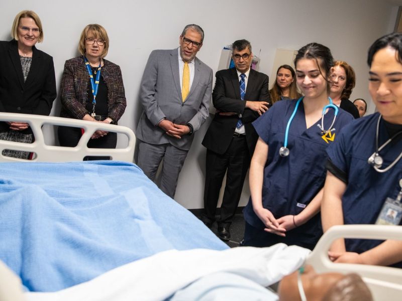 President Lachemi and guests with nursing students in a clinical room with a training mannequin in a hospital bed