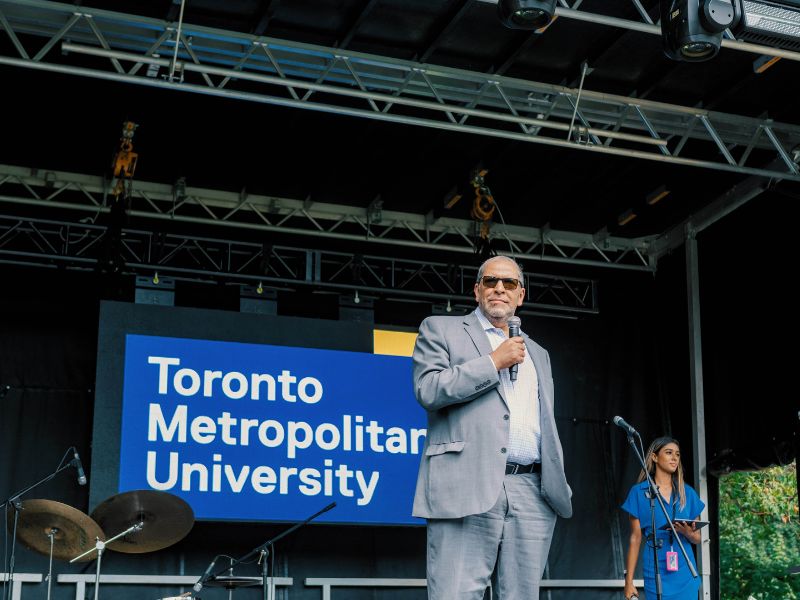 President Lachemi stands on stage at orientation with a microphone in his hand and the TMU logo behind him