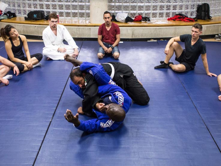 Two men practice Brazilian Jiu Jitsu on a blue mat surrounded by their class.