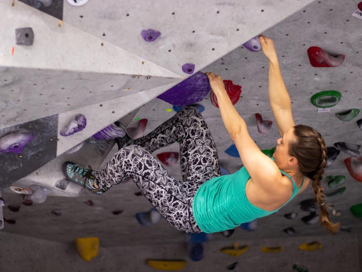 A woman in a green tank top and grey patterned pants climbs an indoor rock climbing wall.