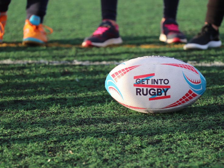 The foreground shows a rugby ball with text that writes, "Get Into Rugby" and players feet in running shoes are in the background.