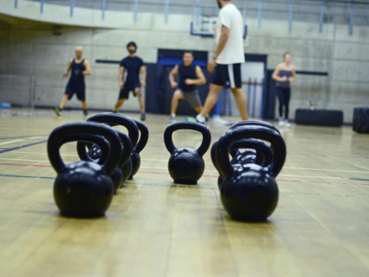 A group of black kettlebells in the foreground and participants warming up in the background.
