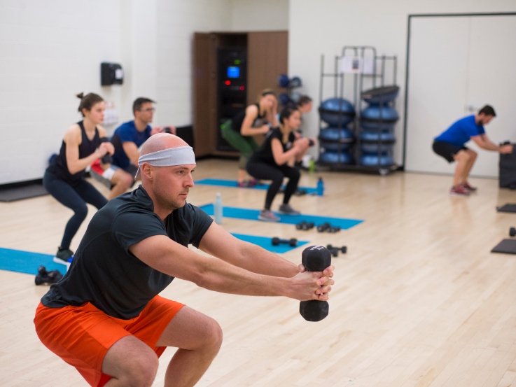 Fitness class participants in the MAC topline studio exercise doing lunges while holding a dumbbell with arms stretched in front of them. 