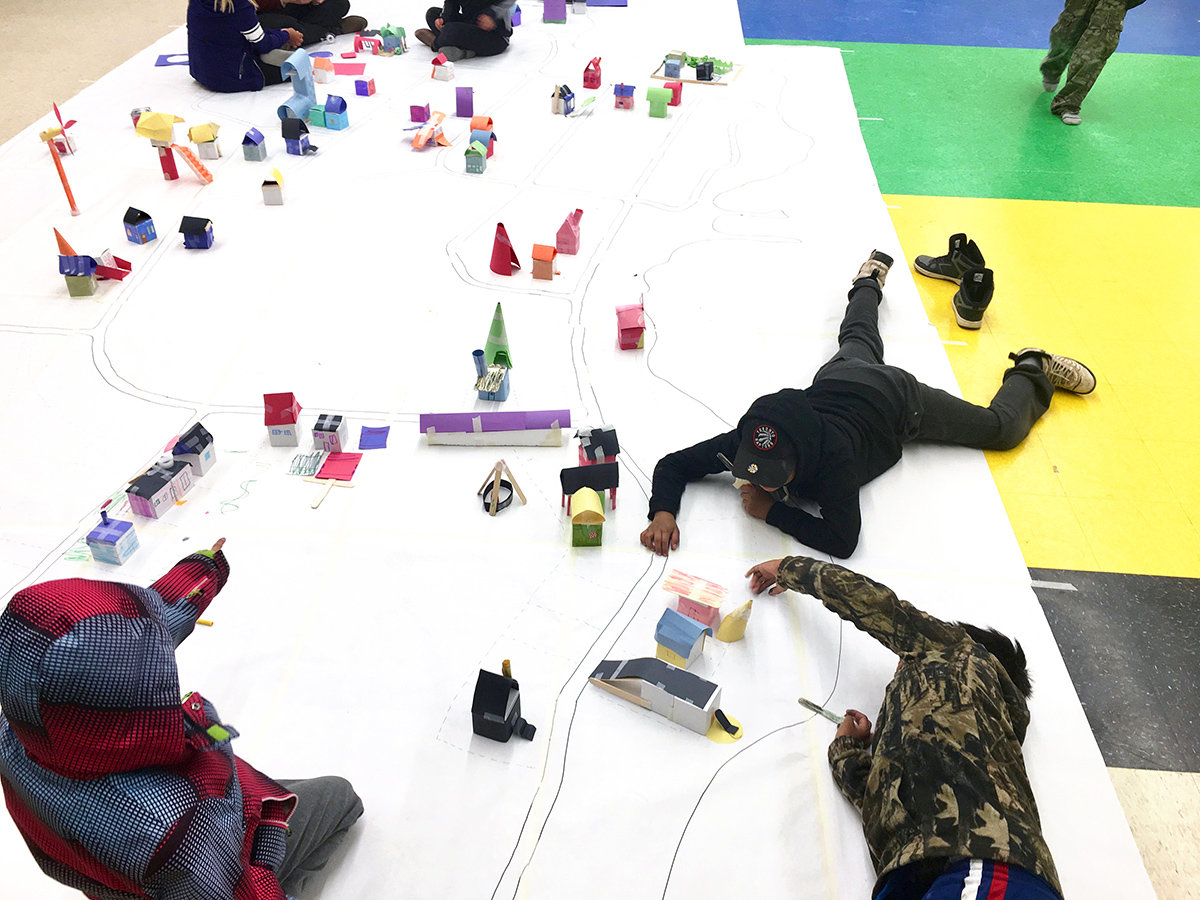 Children lie on top of a miniature city map with small construction paper buildings.