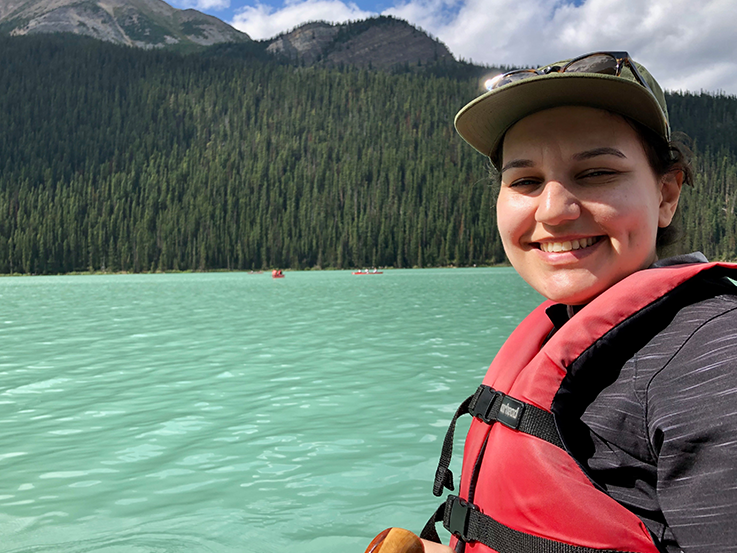  Jessica Machado, sitting in a canoe on a lake, smiling and wearing a red life jacket, with mountains and water in the background. 