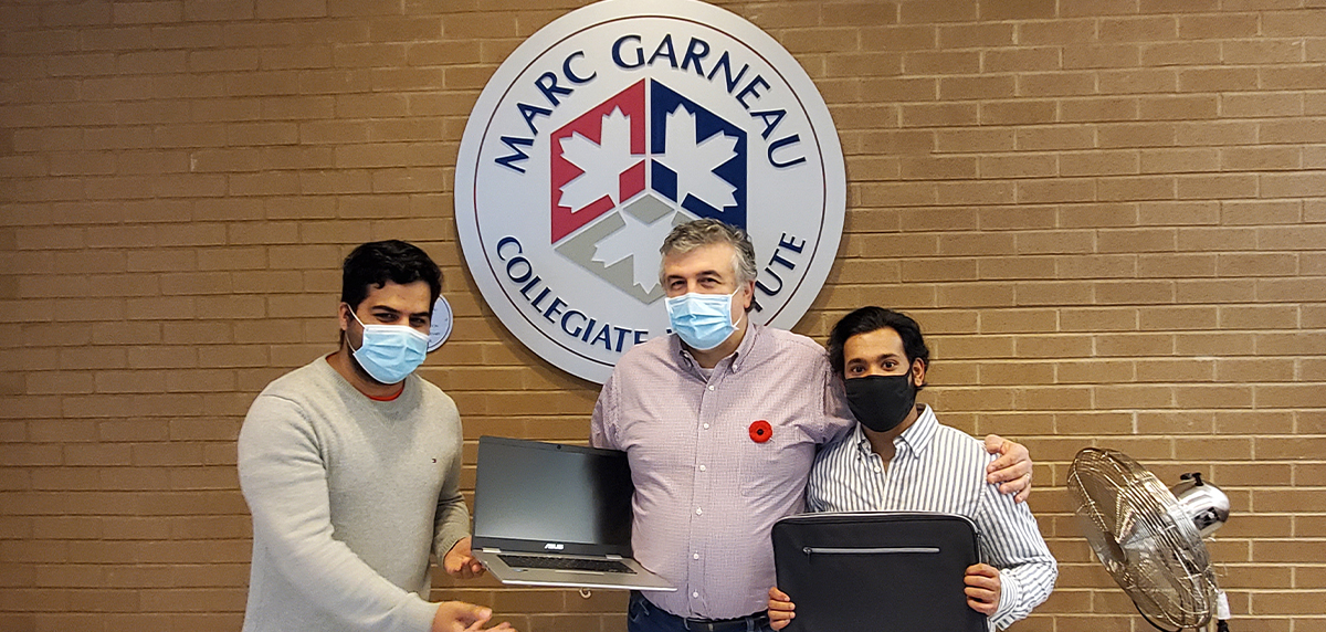 Three men wearing face masks are standing together holding two laptops. Behind them is a red brick wall and a circular sign that reads "MARC GARNEAU COLLEGIATE INSTITUTE."