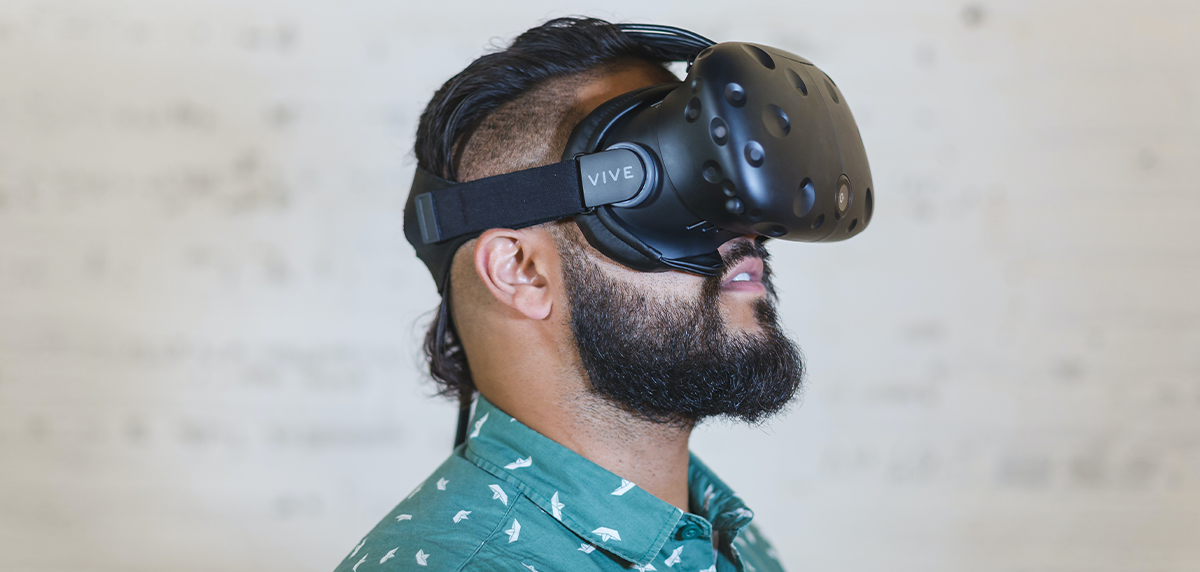 A man with a beard and a mohawk in a light blue shirt with white dots, wearing virtual reality glasses. 