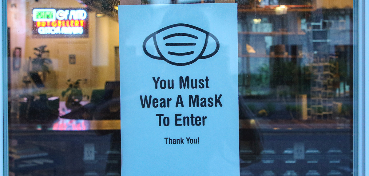 A white sign on the window of a restaurant. The sign has a black and white drawing of a face mask. Under the face mask is the text "You Must Wear A Mask To Enter Thank You!"