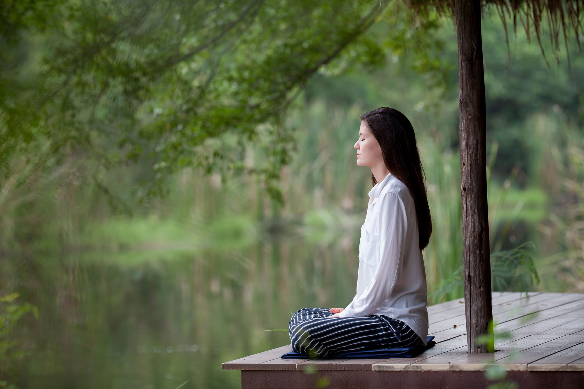 A woman sits on a small porch by the water, legs crossed, the green of the world around her meditative and serene as she sits quietly, reflecting.