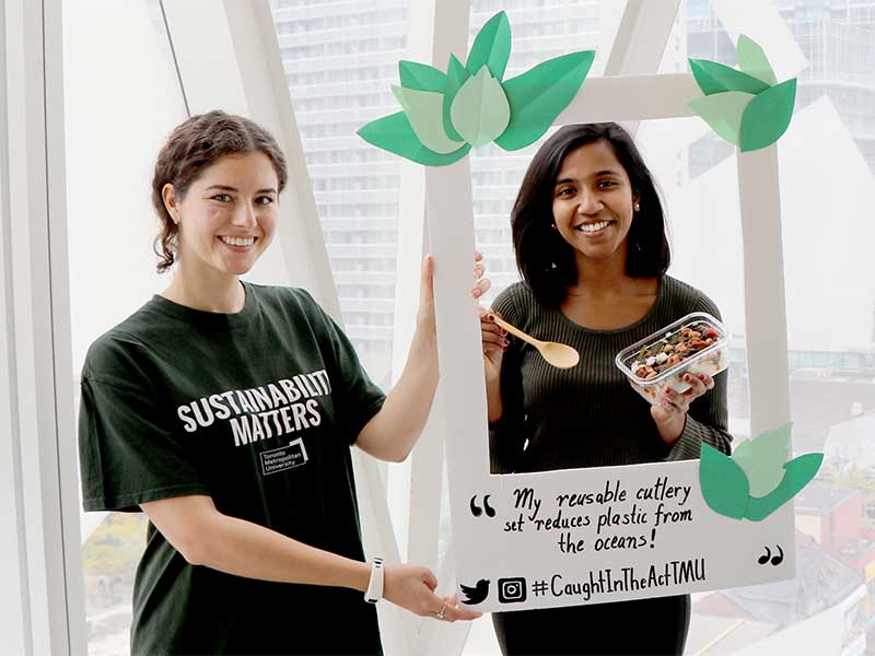 A Sustainability Ambassador holds a decorative, paper frame in front of a student for a social media photo opt that highlights the students good practice of eating lunch with reusable cutlery.