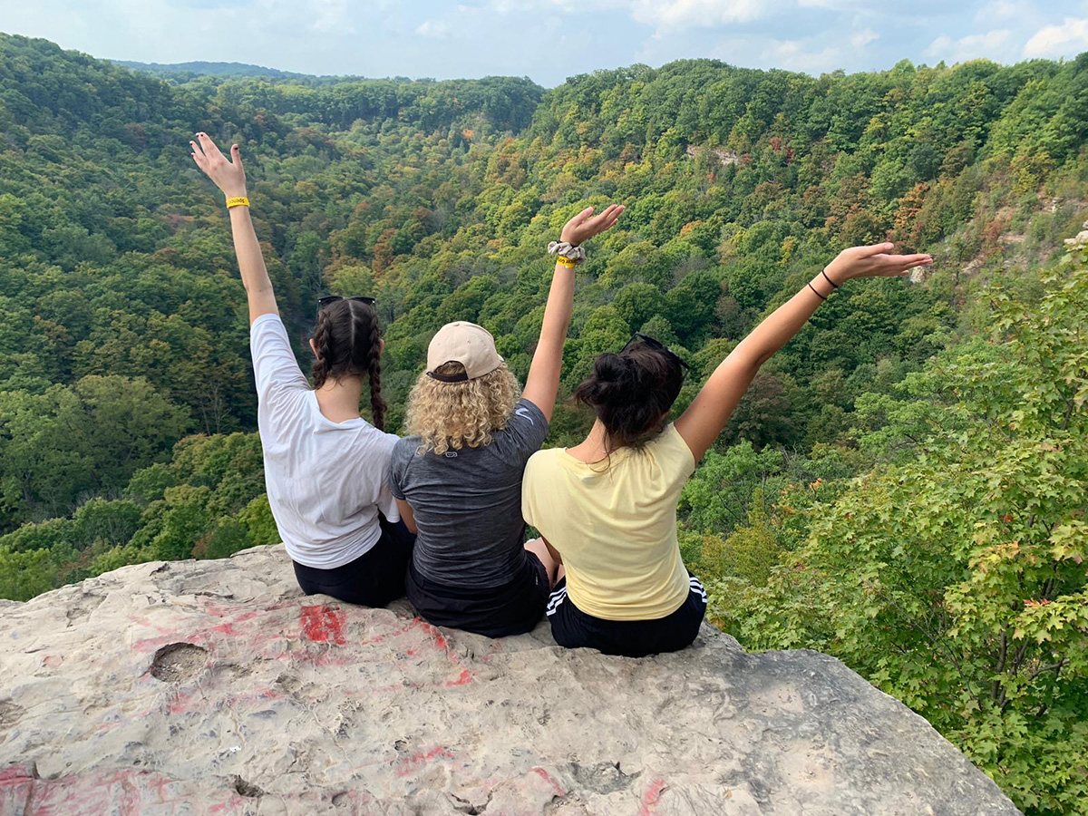 three individuals with hands up in the air while sitting on a rock looking at a lush green forest