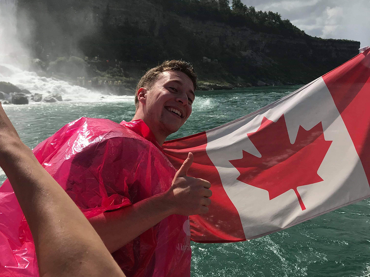 Student smiling at camera with thumbs up and holding the red and white Canadian flag with the water mist in the background