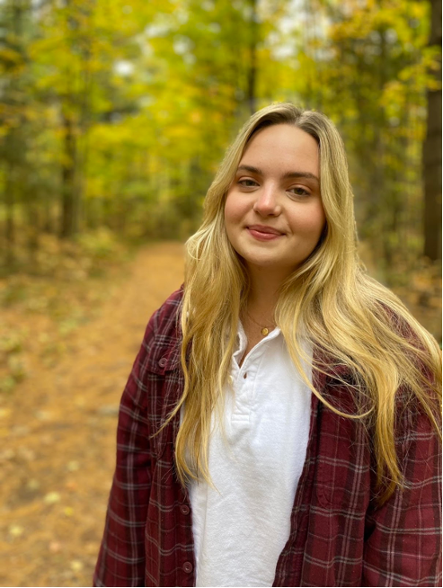 Portrait of Emily Duncan. Emily is smiling in the woods wearing a white and red shirt. 