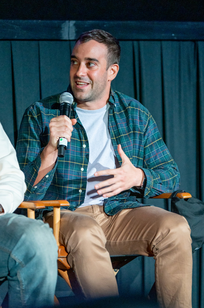 Photograph of Michael Grassi at a panel speaking into a microphone. Michael wears a blue button down and beige pants