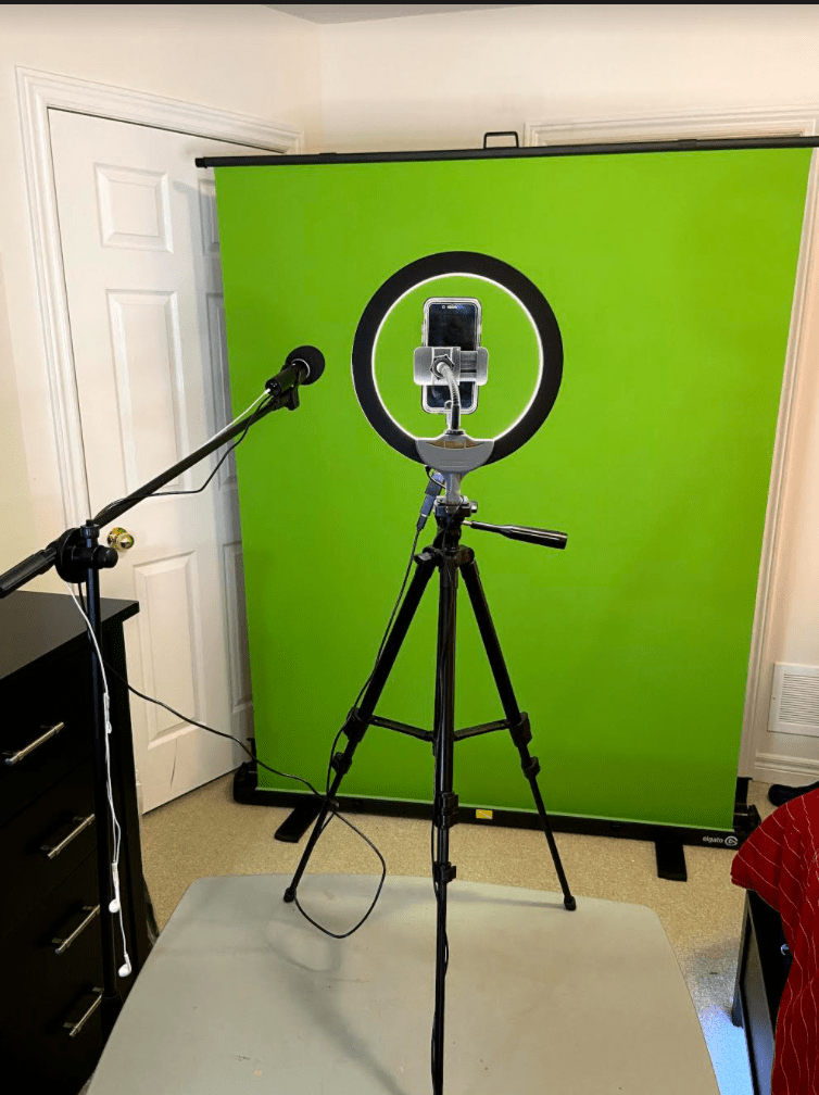 Photograph shows Dimuccio’s filming set-up. In front of a green screen is his iPhone on a tripod along with a ring light and a microphone