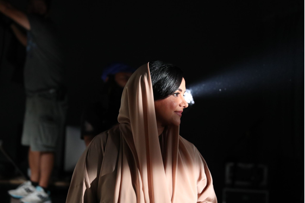 Nayla Al Khaja stands in front of a production light and looks to the right, smiling. She is wearing light pink and is on set.