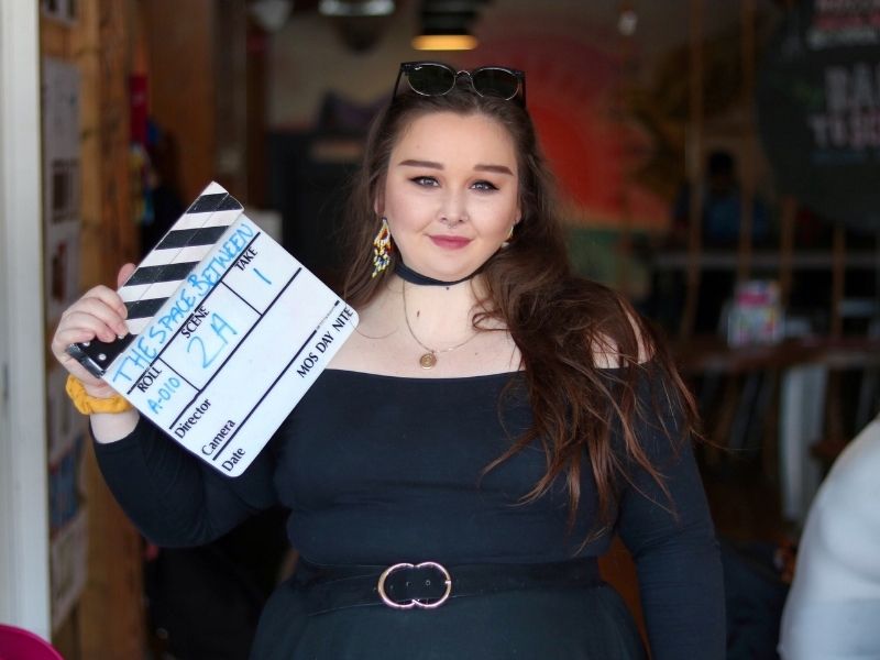 Karly standing in a doorway with film clapper. She wears a black belted dress and beaded earrings.