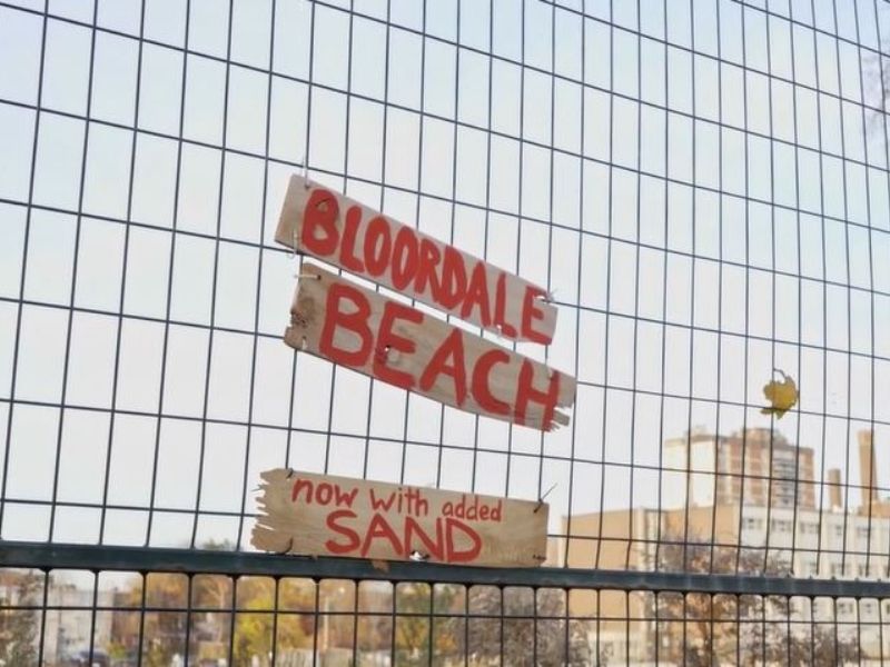 Wooden signs affixed to fence read 'Bloordale Beach' and 'now with added sand' in red