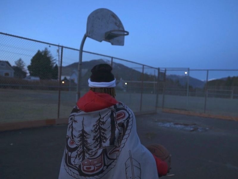 Josiah from the back on a basketball court in the early evening wearing an Indigenous patterned blanket over his shoulders