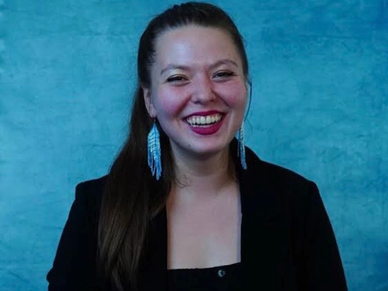Sarah Elaine McLay wearing a black blazer and blue beaded earrings standing in front of a blue background