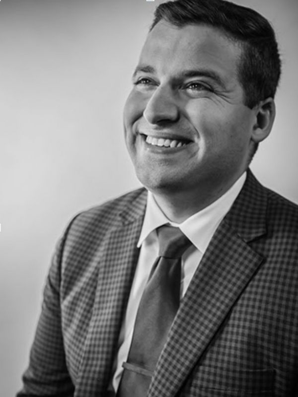 Black and white image of Derek Ryder in a checked blazer and tie, smiling