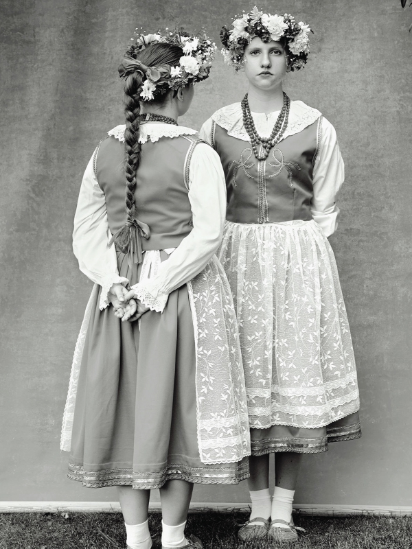 A black and white image of two girls standing wearing Victorian style dresses and flower crows. One girl faces the camera and the other has her back turned
