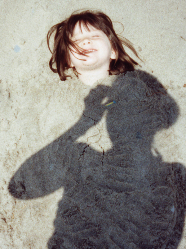 An image of a little girl laughing buried in sand with her mother's shadow taking a picture overtop