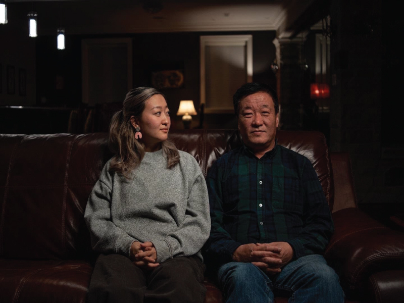 A man and woman sit on a brown couch in a dark room. The woman is looking at the man