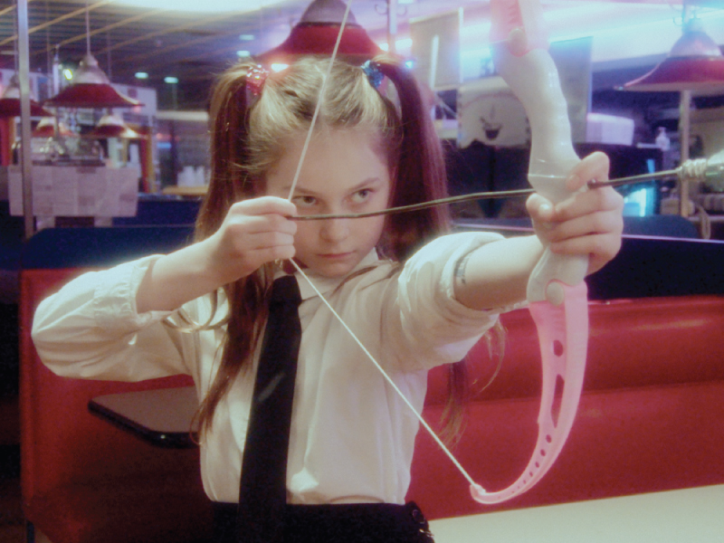 Film still from ‘Sugar Baby’.  A young girl stands in a diner holding a pink plastic bow and arrow