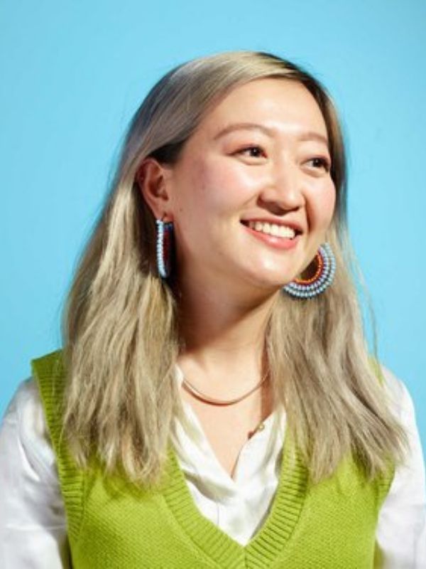 Young Asian woman with blonde hair wears white collared shirt, green knit vest and beaded hoop earrings